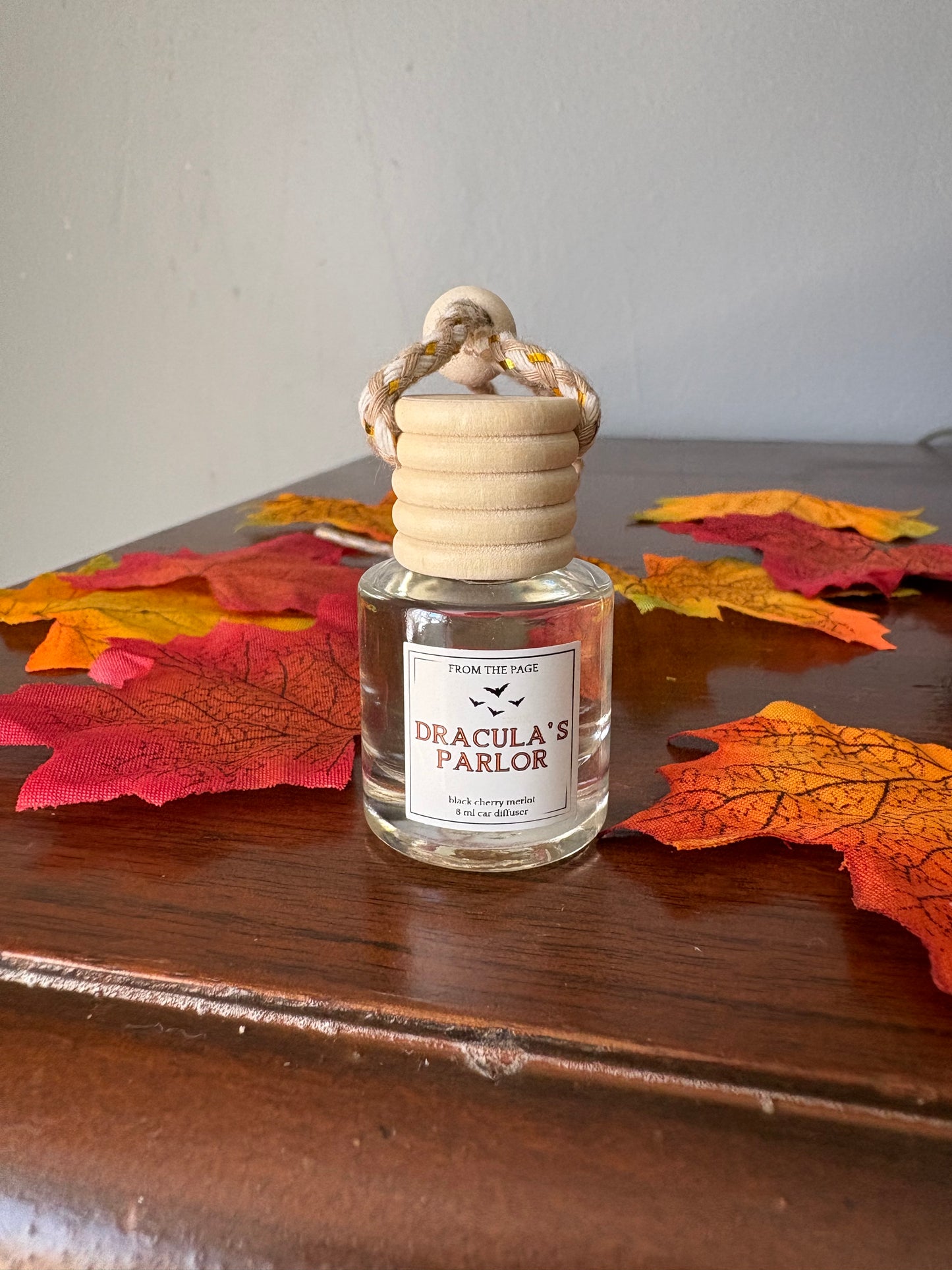 A small clear glass bottle with a beige ribbed cap and hanging loop, labeled 'Dracula's Parlor' by From The Page, placed on a wooden surface with red and orange autumn leaves around it.