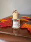 A small clear glass bottle with a beige ribbed cap and hanging loop, labeled 'Dracula's Parlor' by From The Page, placed on a wooden surface with red and orange autumn leaves around it.