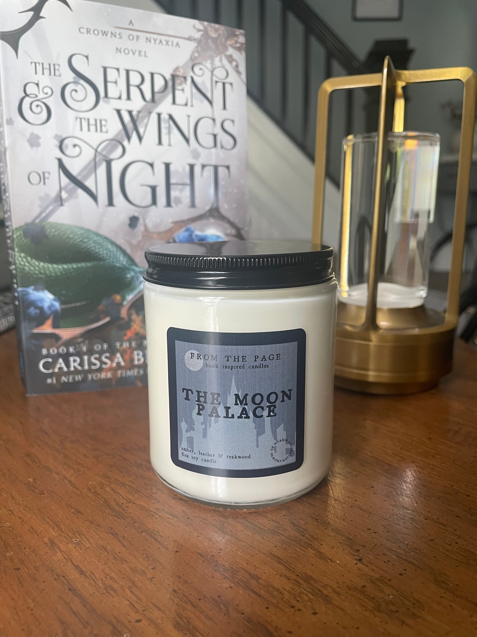Candle with a label on a wooden surface next to a book titled 'Serpent and the Wings of Night'.