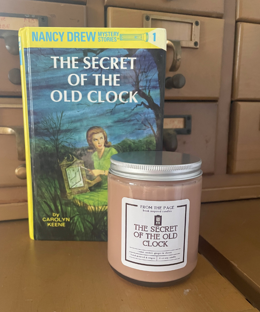Candle titled 'The Secret of the Old Clock' next to a Nancy Drew book on a wooden surface.