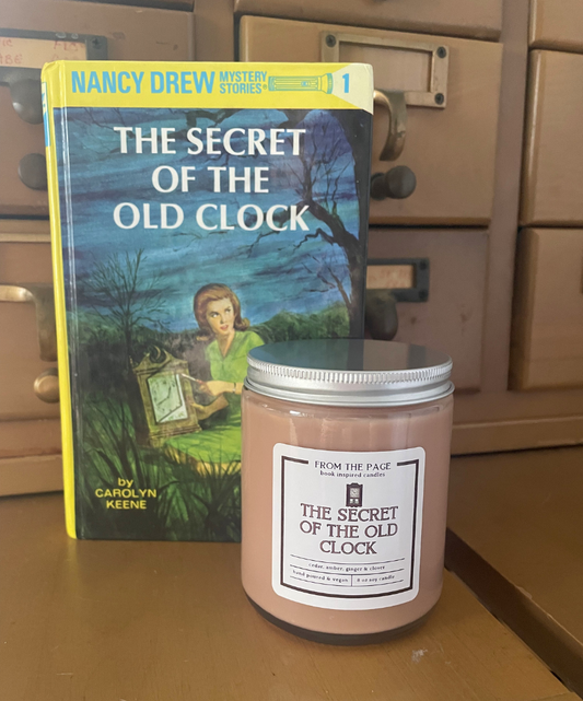 Candle titled 'The Secret of the Old Clock' next to a Nancy Drew book on a wooden surface.