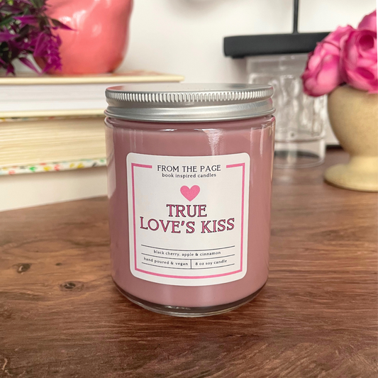 a pink candle with the label "true love's kiss" sitting on a wooden board with a stack of books in the background