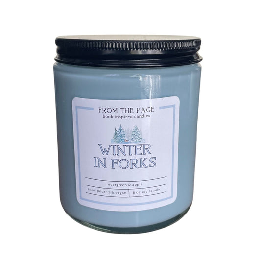 a light blue candle with a black lid with the label reading Winter in Forks. A small photo of a snowy forest is on top, with the scent reading evergreen and apple