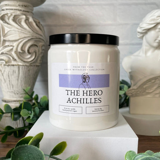 white candle jar with the label "The Hero Achilles" and a black lid. Green leaves and Greek statue in the background