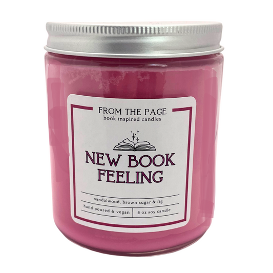 A burgundy soy wax candle in a clear jar with a metal lid, labeled 'NEW BOOK FEELING' with the From The Page logo, and scents listed as sandalwood, brown sugar & fig.